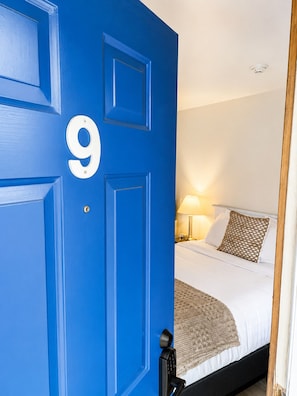 A room with a blue door marked with the number 9, a bed with white bedding, a lamp, and a textured throw blanket.