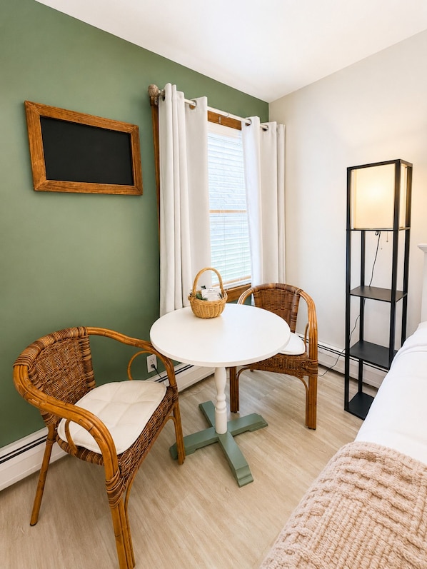 A room with a round table, two wicker chairs, a basket, a floor lamp, and a bed.