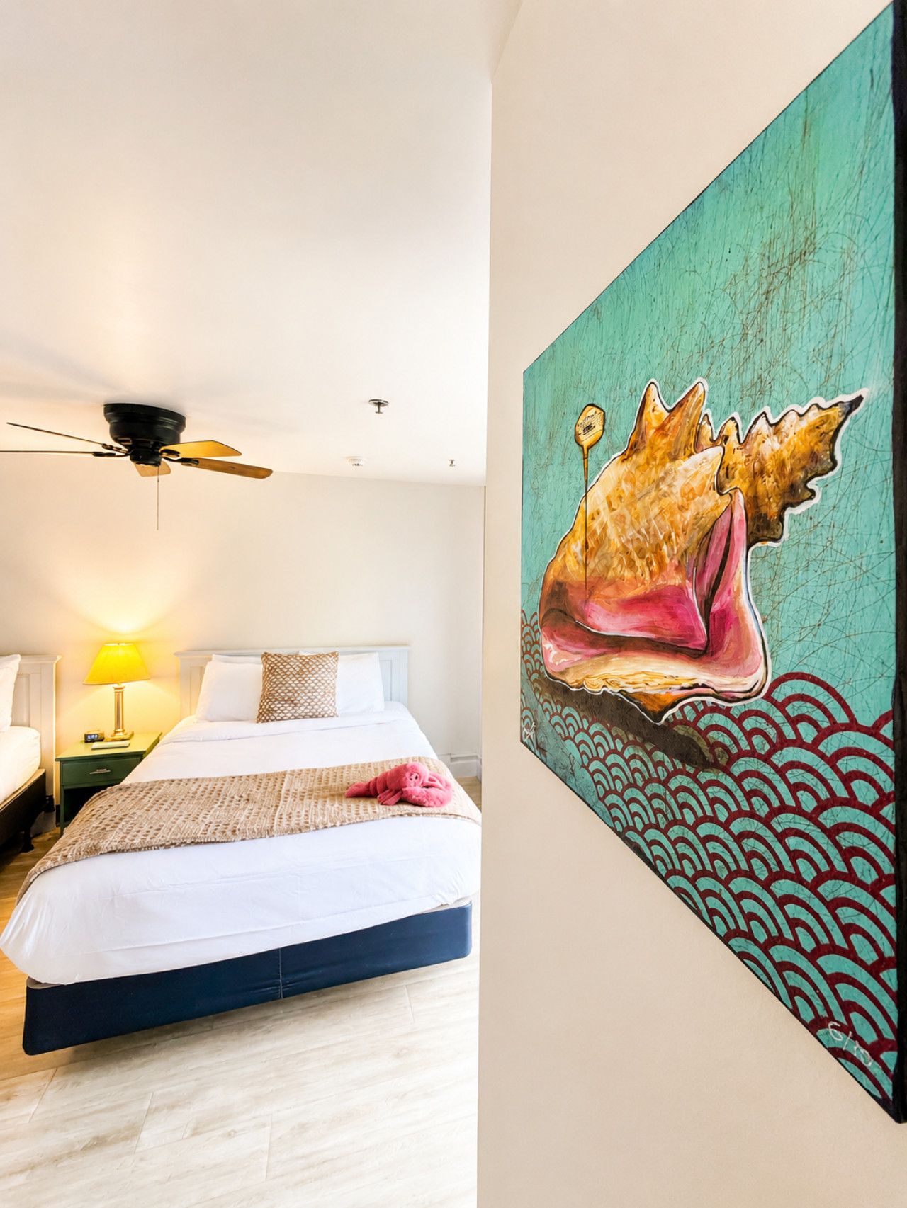 A hotel room with a bed, a ceiling fan, a nightstand with a lamp, and a large painting of a shell on the wall.