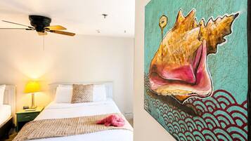 A hotel room with a bed, a ceiling fan, a nightstand with a lamp, and a large painting of a shell on the wall.