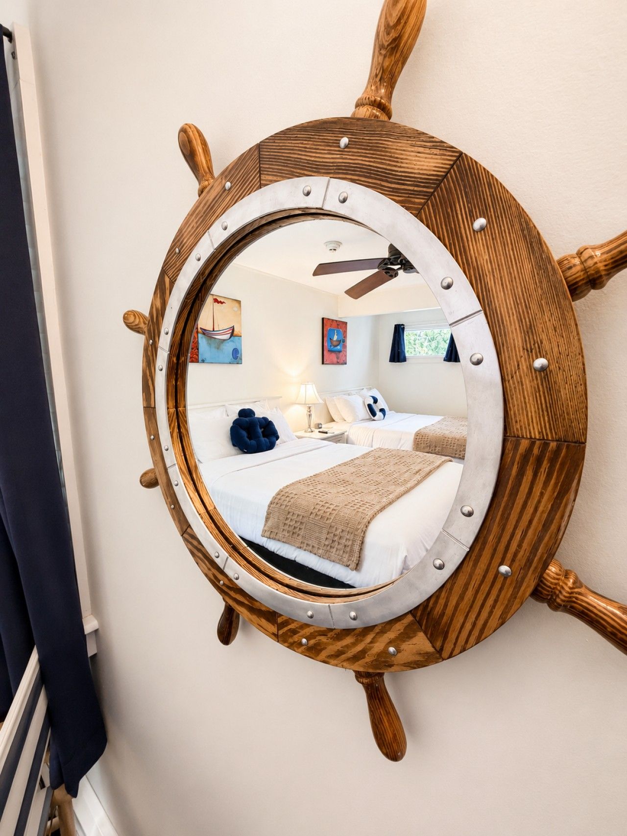 A round mirror with a ship's wheel design, reflecting a bedroom with a bed, pillows, and a lamp.