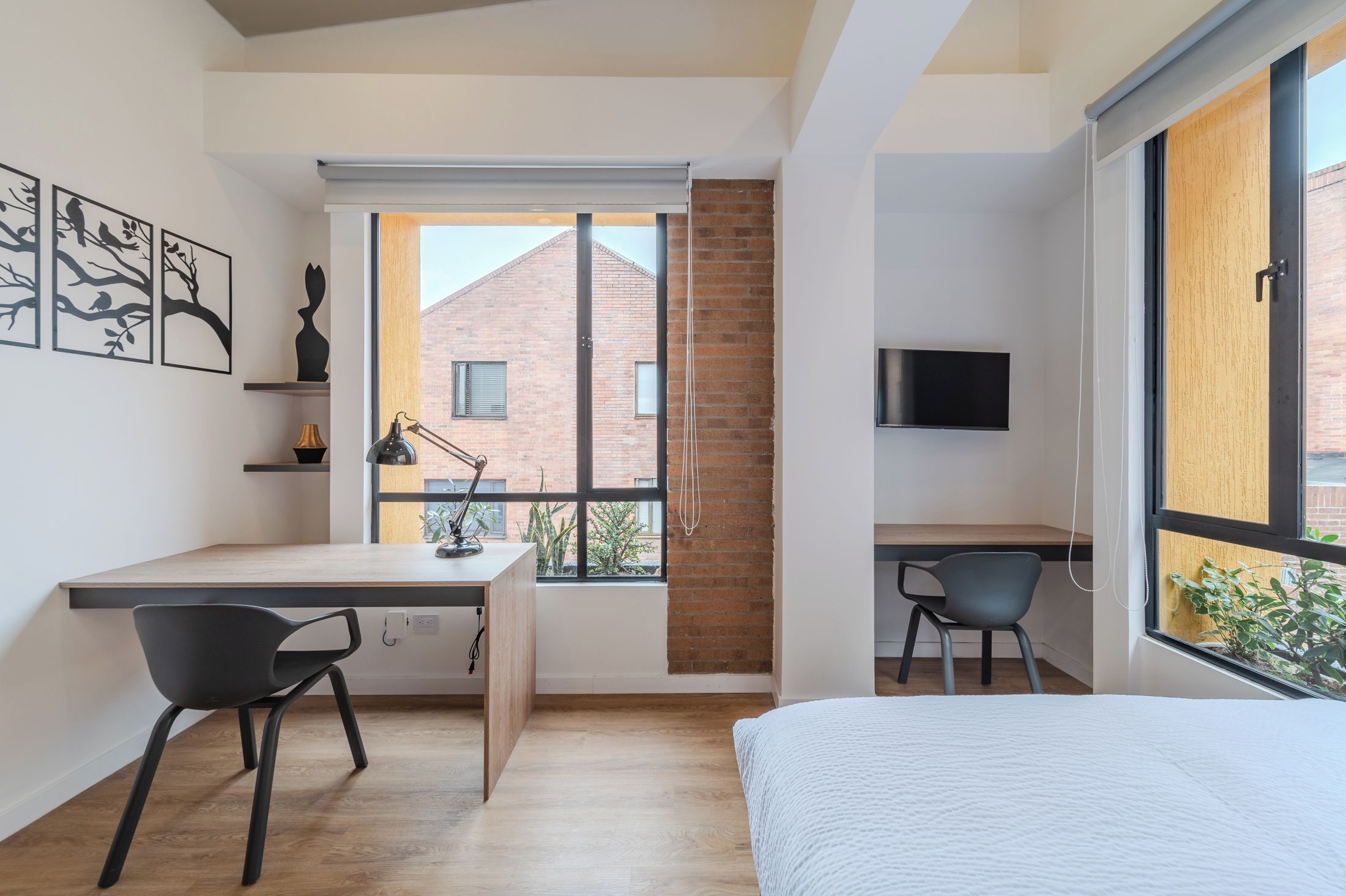Une chambre moderne avec un grand lit, un bureau et une chaise, un téléviseur fixé au mur et une fenêtre donnant sur un immeuble en briques.