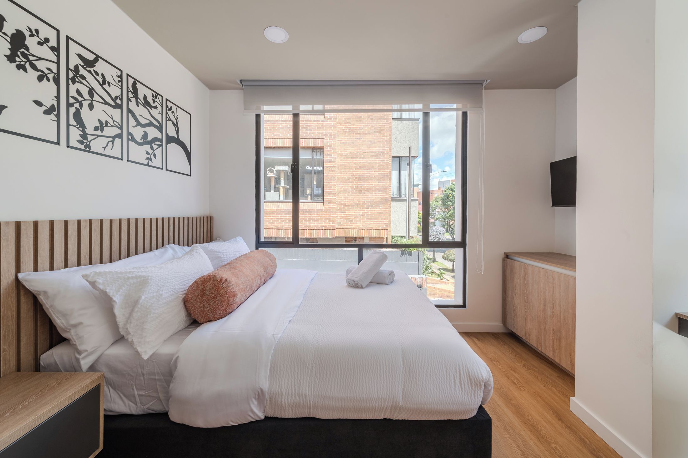 A modern bedroom with a large bed, a wooden headboard, a window with a view of buildings, and a flat-screen TV on the wall.