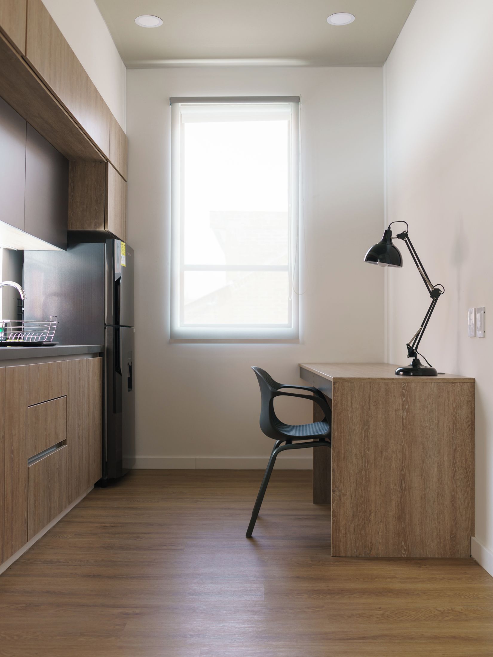 Een kleine keuken met houten kastjes, een inbouwkoelkast, een raam en een bureau met stoel.