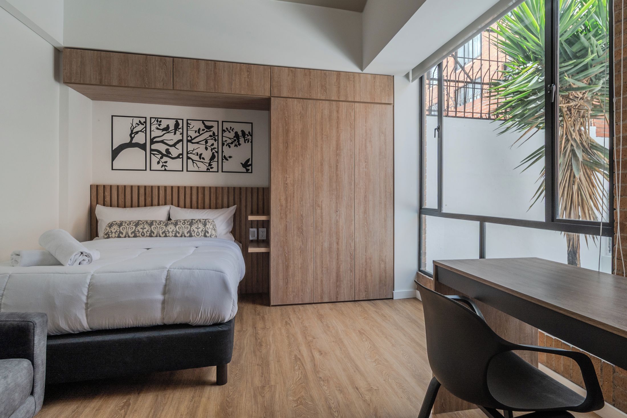 A bedroom with a bed, a desk, and a window with a view of a building and plants.