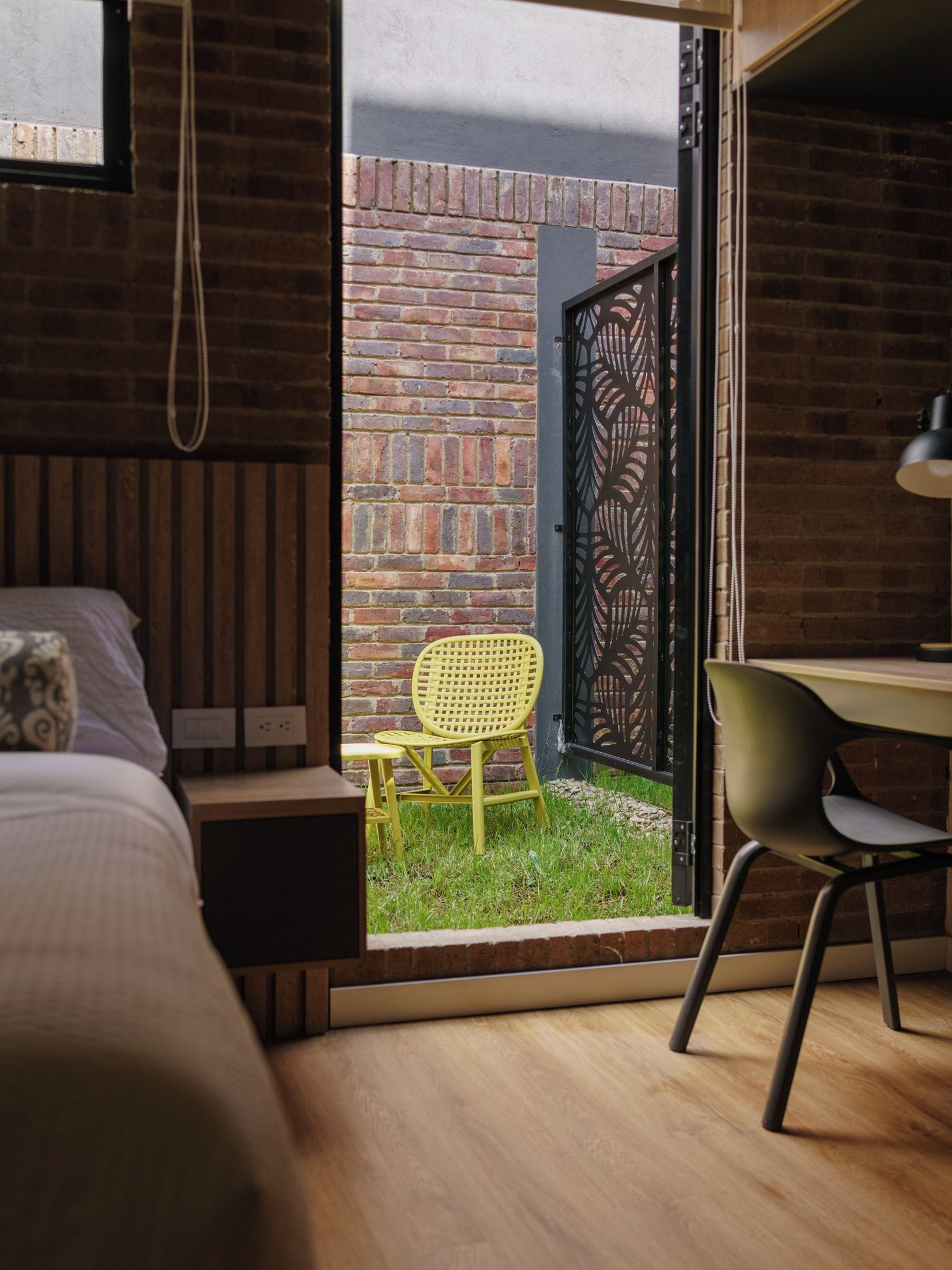 Quarto com cama, escrivaninha e cadeira. Tem uma janela com vista para uma parede de tijolos e área gramada.