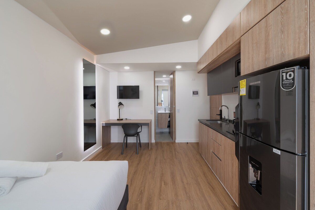 Habitación de hotel pequeña con cama, escritorio, silla, kitchenette con refrigerador y televisor montado en la pared.