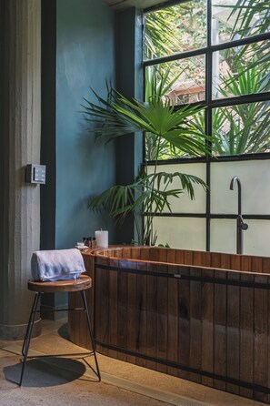 Un baño con una bañera de madera, una planta en maceta, una toalla sobre un taburete y una ventana con vista a follaje.