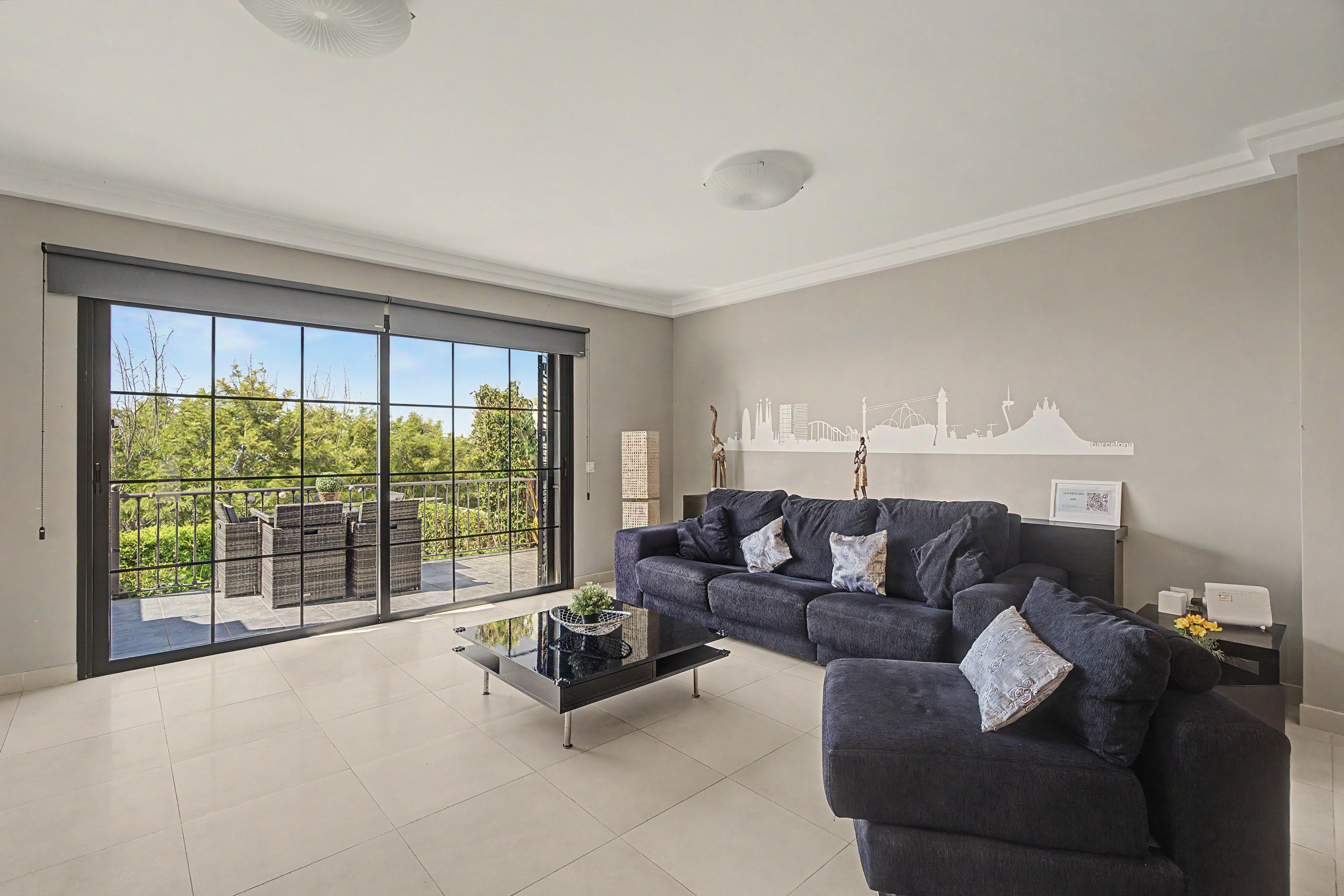 A spacious living room with a large sliding glass door leading to an outdoor balcony, a dark-colored sofa set, and a modern coffee table.