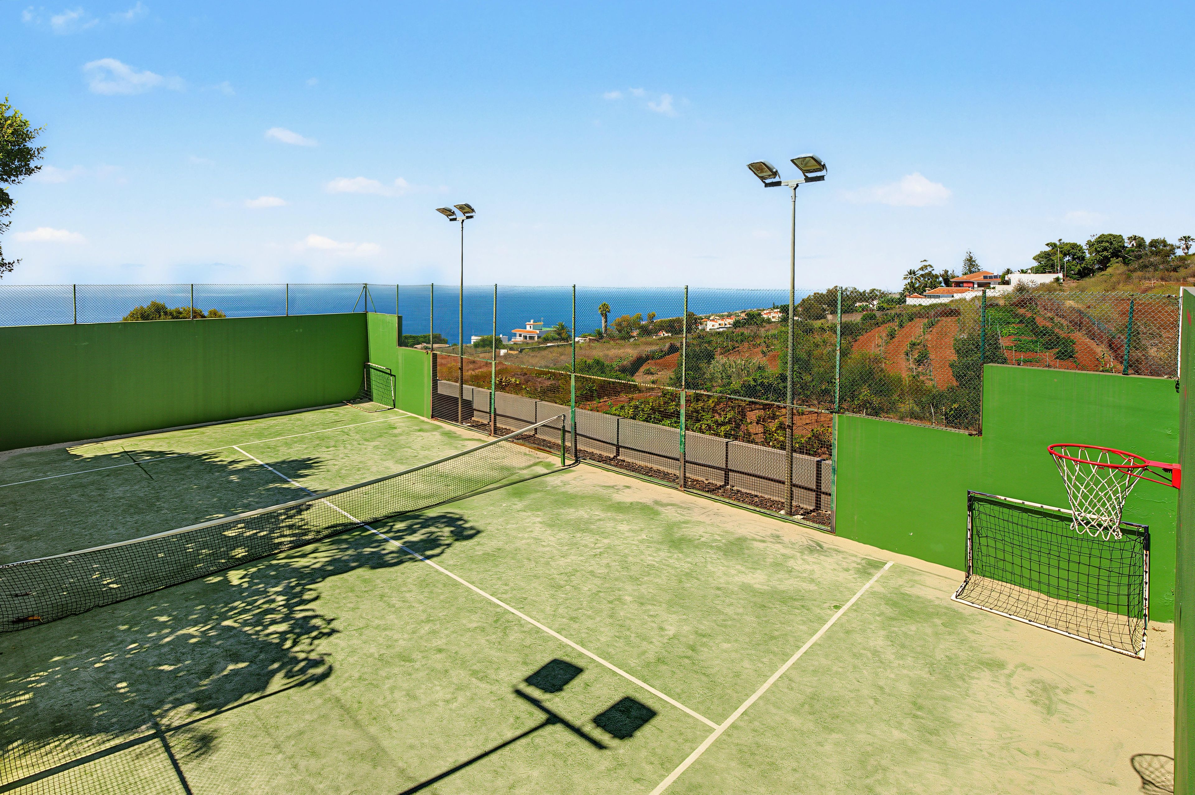 Ein Tennisplatz mit Netz, umgeben von einem grünen Zaun, mit Blick auf eine Küstenlandschaft mit Häusern und Bäumen.