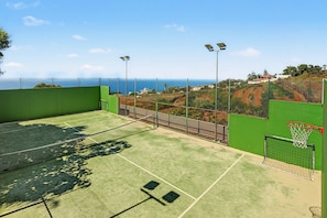 Una cancha de tenis con red, rodeada por una valla verde y con vistas a una zona costera con casas y árboles.
