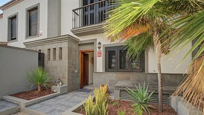 Um edifício residencial com entrada fechada por portão, uma varanda com guarda-corpo e uma pequena área de jardim com plantas e uma palmeira.