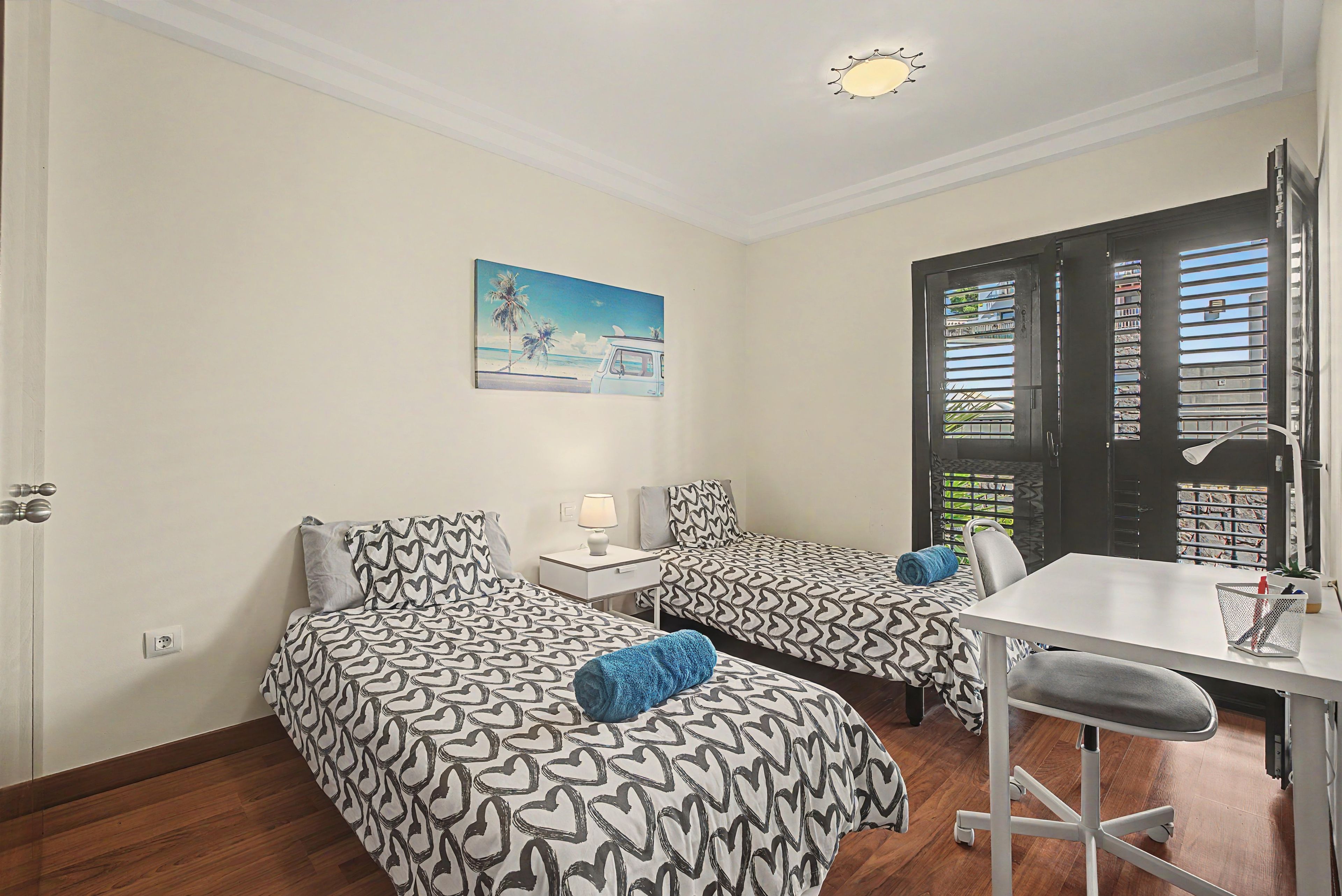 A room with two beds, a desk, a chair, and a window with blinds.