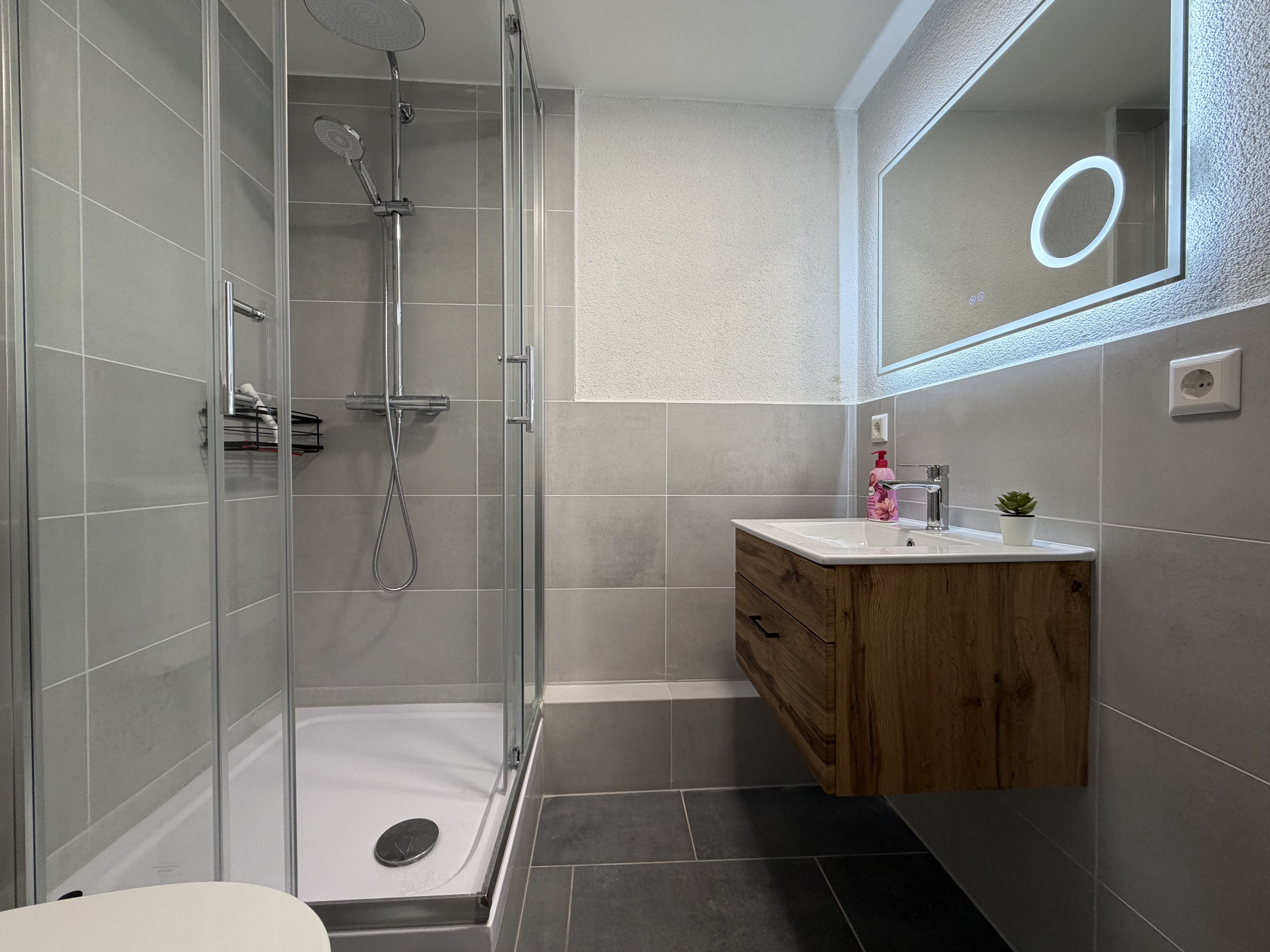 A modern bathroom with a glass-enclosed shower, a wooden vanity, and a large mirror with a circular light.