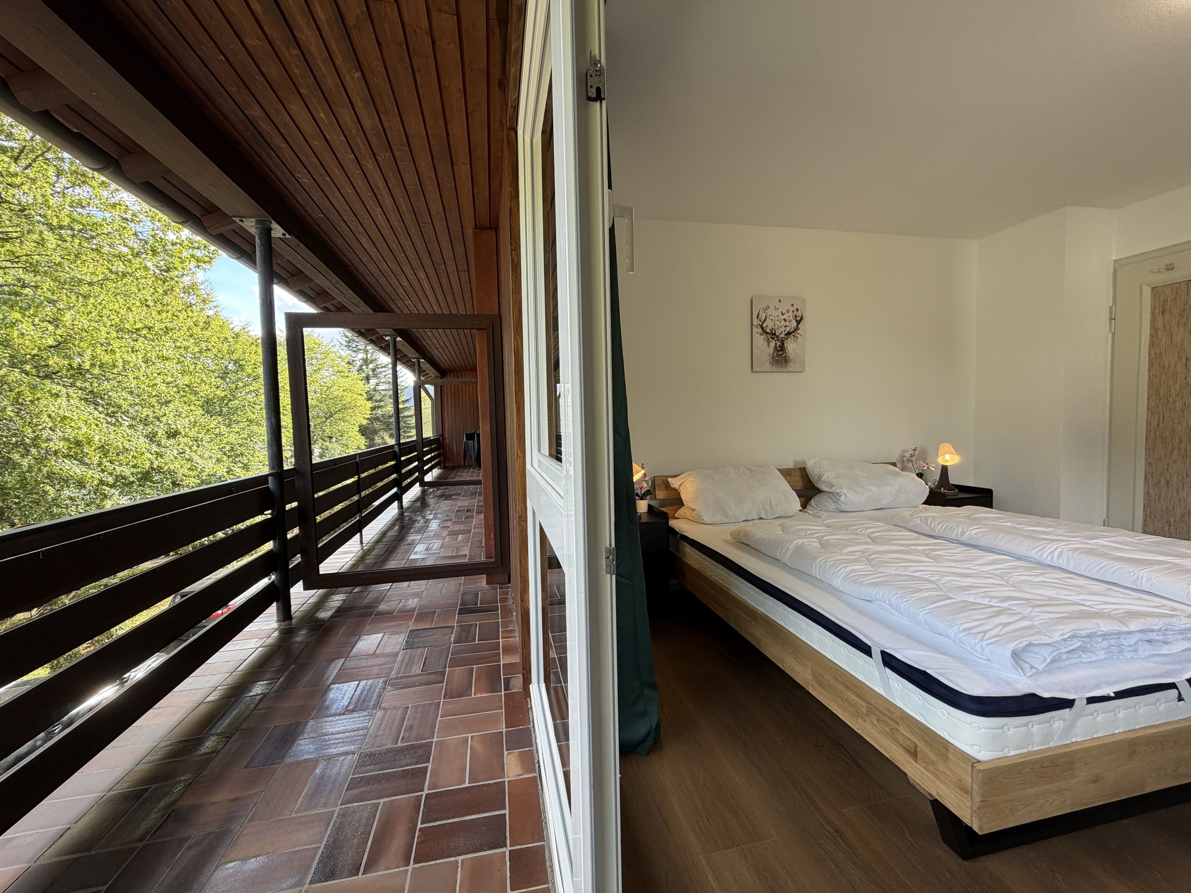 A bedroom with a wooden bed, a nightstand with a lamp, and a view of trees through an open window.
