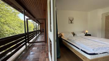 A bedroom with a wooden bed, a nightstand with a lamp, and a view of trees through an open window.