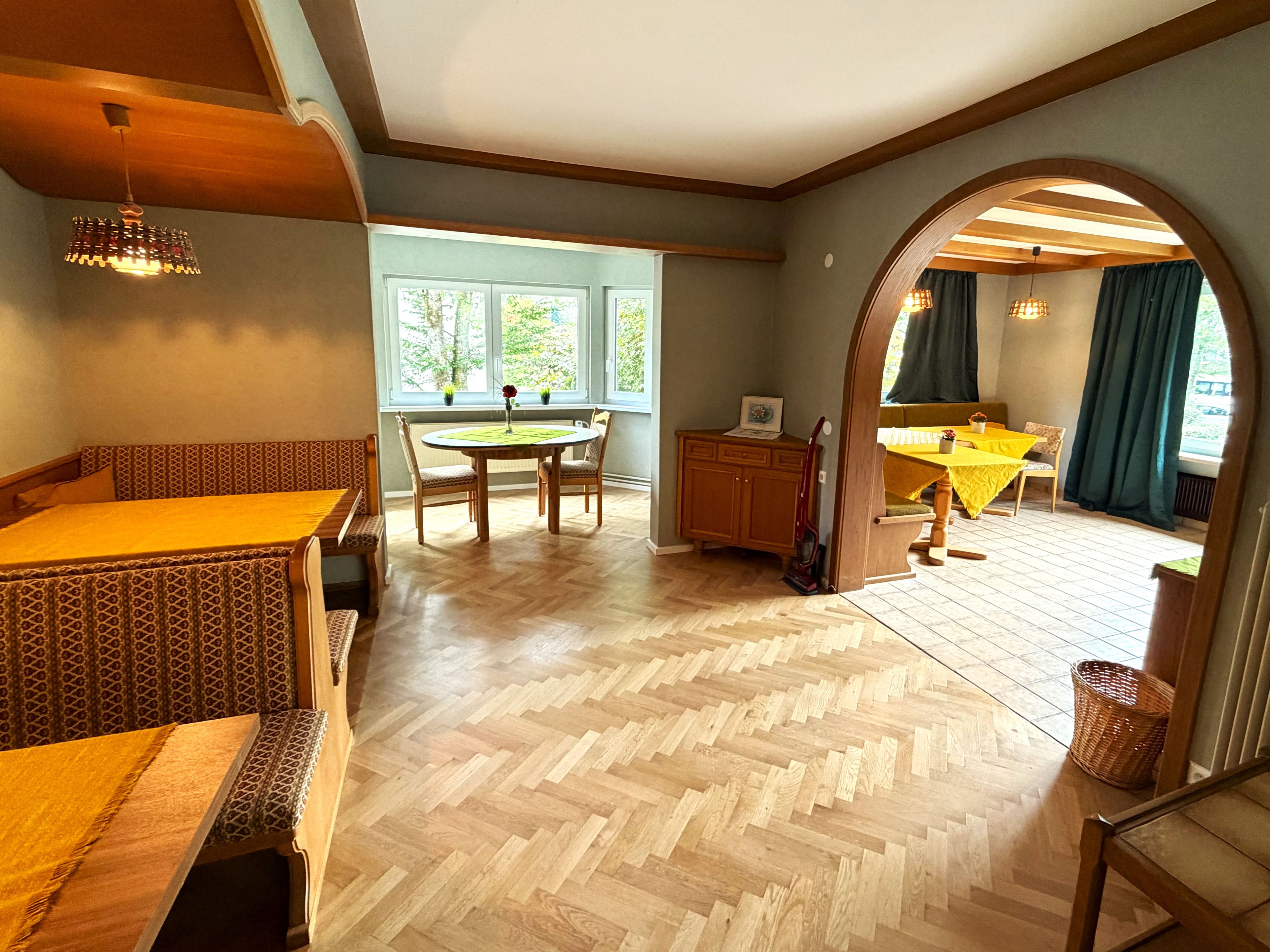 A room with a herringbone floor, a dining table with chairs, a bench, a small cabinet, and a window with a view of trees.