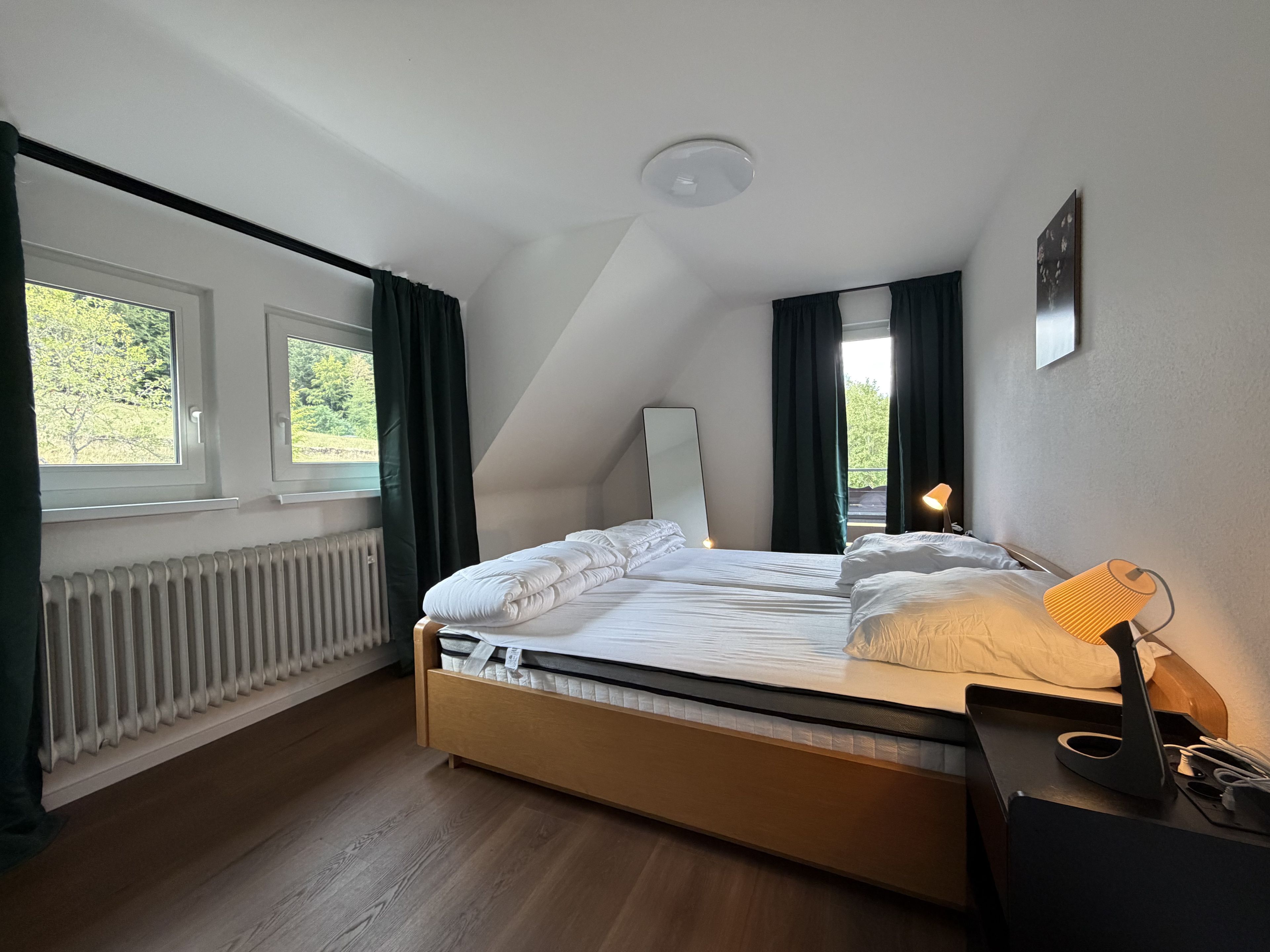 A bedroom with a bed, a bedside lamp, a radiator, and windows with curtains.
