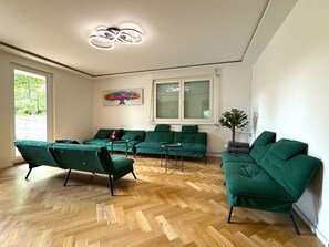 Un salon moderne avec des canapés verts, un parquet en bois disposé en chevrons, un tableau au mur et une fenêtre avec des stores.