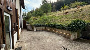 Une cour en gravier avec un bâtiment en bois, un mur de pierre incurvé et une colline plantée d’arbres, où se trouve une vache.