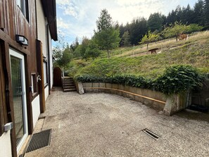 A gravel courtyard with a wooden building, a curved stone wall, and a hillside with trees and a cow.