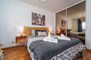Ein Schlafzimmer mit einem großen Bett, einem hölzernen Kopfteil, einem Nachttisch mit Lampe, einem Bild an der Wand und einem Spiegel, der den Raum widerspiegelt.