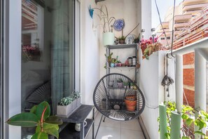 Ein kleiner Balkon mit einem Stuhl, Topfpflanzen und einem Metallregal mit diversen Gegenständen.