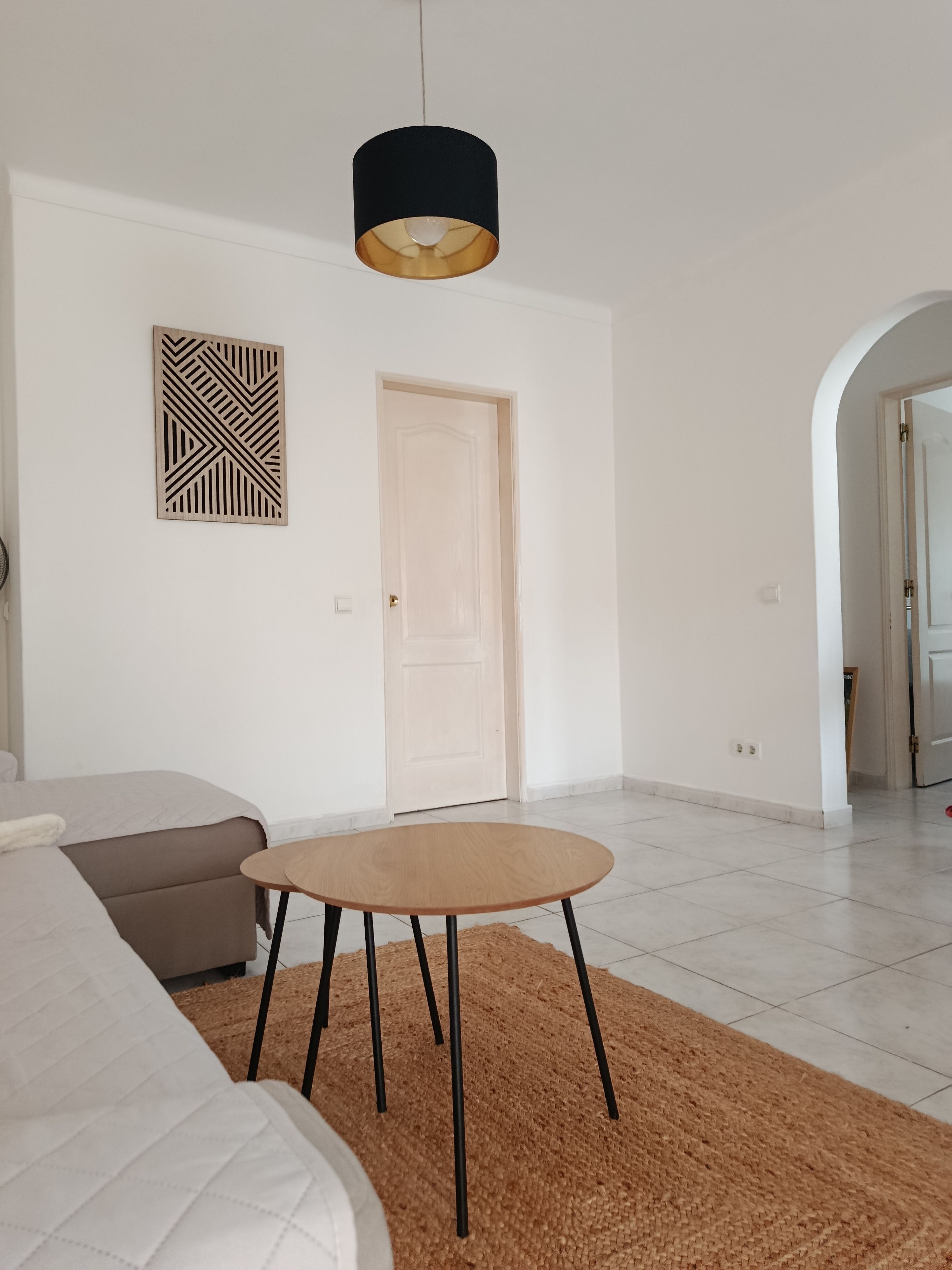 A modern living room with a round wooden table, a beige sofa, a black hanging lamp, and a geometric wall art piece.