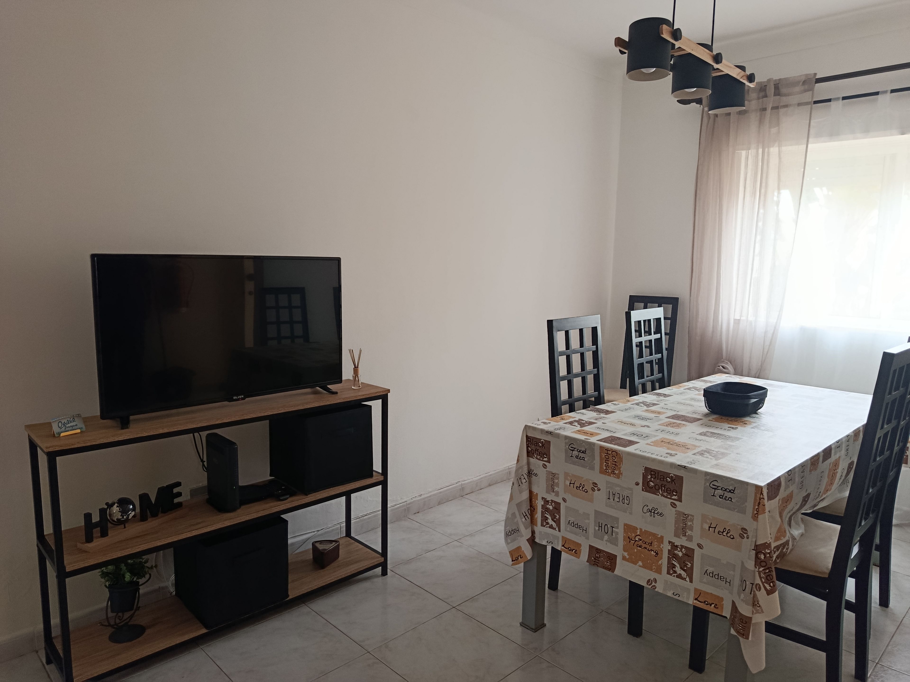 Un saló modern amb un televisió de pantalla plana sobre un suport de fusta, una taula de menjador amb cadires i una finestra amb cortines lleugeres.