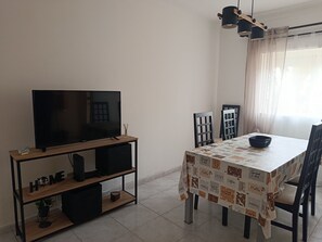 Een moderne woonkamer met een flatscreen televisie op een houten standaard, een eettafel met stoelen, en een raam met dunne gordijnen.