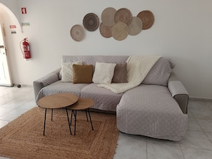 Een woonkamer met een grijze bank, twee ronde bijzettafeltjes, een bruin vloerkleed en een brandblusser aan de muur.