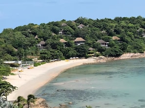Una zona costera con una playa arenosa, agua cristalina y una colina con varios edificios.