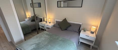 A bedroom with a bed, two bedside tables, a mirror, and a wall-mounted light fixture.