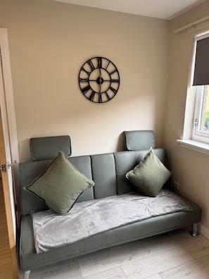 A modern living room with a grey sofa, two green cushions, a grey throw blanket, and a wall-mounted clock. - Newly Renovated 1 Bedroom Flat In Edinburgh - Sleeps 4 People (Edinburgh)