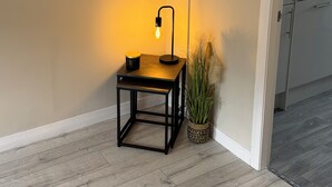 Ett hörnrum med ett litet bord, en golvlampa, en krukväxt och ett inramat konstverk på väggen.