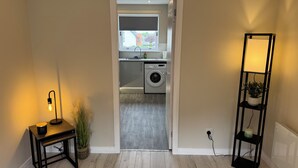 En hall som leder till ett utrymme med tvättmaskin, ett litet bord med en lampa och en hög hylla med en växt.