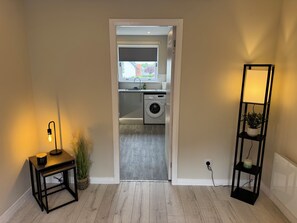 A hallway leading to a laundry area with a washing machine, a small table with a lamp, and a tall shelf with a plant. - Newly Renovated 1 Bedroom Flat In Edinburgh - Sleeps 4 People (Edinburgh)