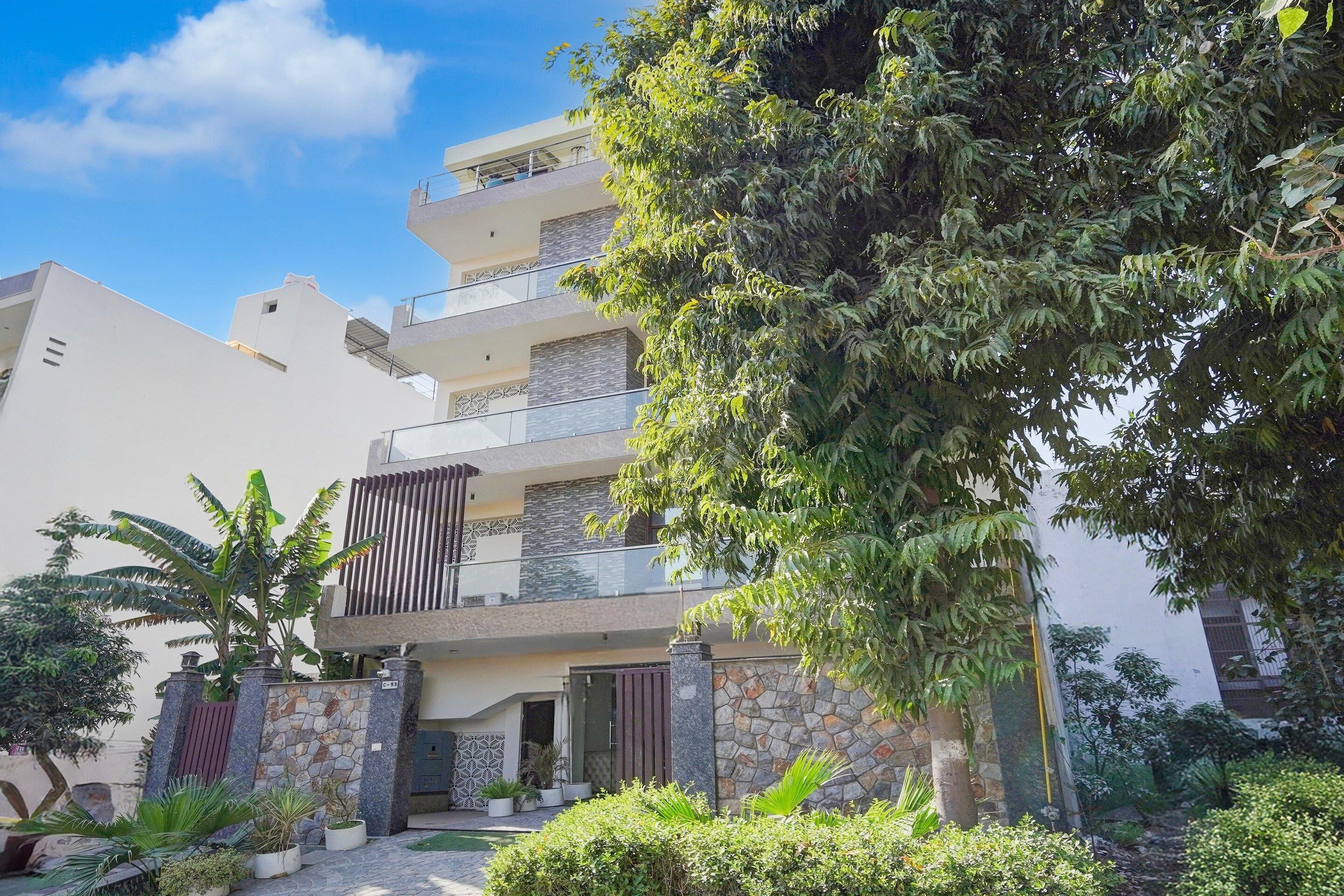 A modern multi-story residential building with a stone facade, a balcony, and a well-maintained garden with various plants.