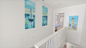 A hallway with white wainscoting and a white railing. There are three paintings on the walls: two of them depict sailboats on a blue water background, and one shows a colorful beach scene with a lifebuoy. - 12B - 2BR $700+ in freebies incl golf, On Beach (Miramar Beach)