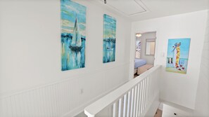 A hallway with white wainscoting and a white railing. There are three paintings on the walls: two of them depict sailboats on a blue water background, and one shows a colorful beach scene with a lifebuoy. - 12B - 2BR $700+ in freebies incl golf, On Beach (Miramar Beach)