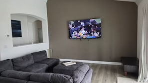 Ein Wohnzimmer mit einem großen Flachbildfernseher an der Wand, einem dunkelgrauen Sofa und einem kleinen Hocker.