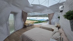 Ein modernes, geometrisches Interieur mit einem großen Bett, einem kleinen Tisch und Blick auf ein grünes Feld durch eine transparente Decke.
