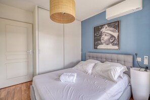 Un dormitori amb un llit, aire condicionat fixat a la paret, un quadre amb marc i una tauleta de nit amb una làmpada.