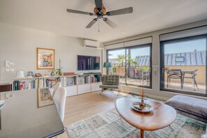 A modern living room with a large window, a ceiling fan, a flat-screen TV, and a bookshelf. - Stunning Flat • AC & Parking in Bordeaux (Bordeaux)