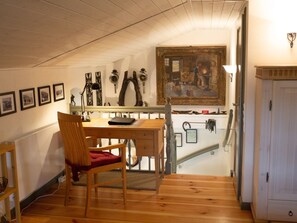 A cozy room with a wooden desk, a chair with a red cushion, a painting on the wall, and a staircase leading to another level.