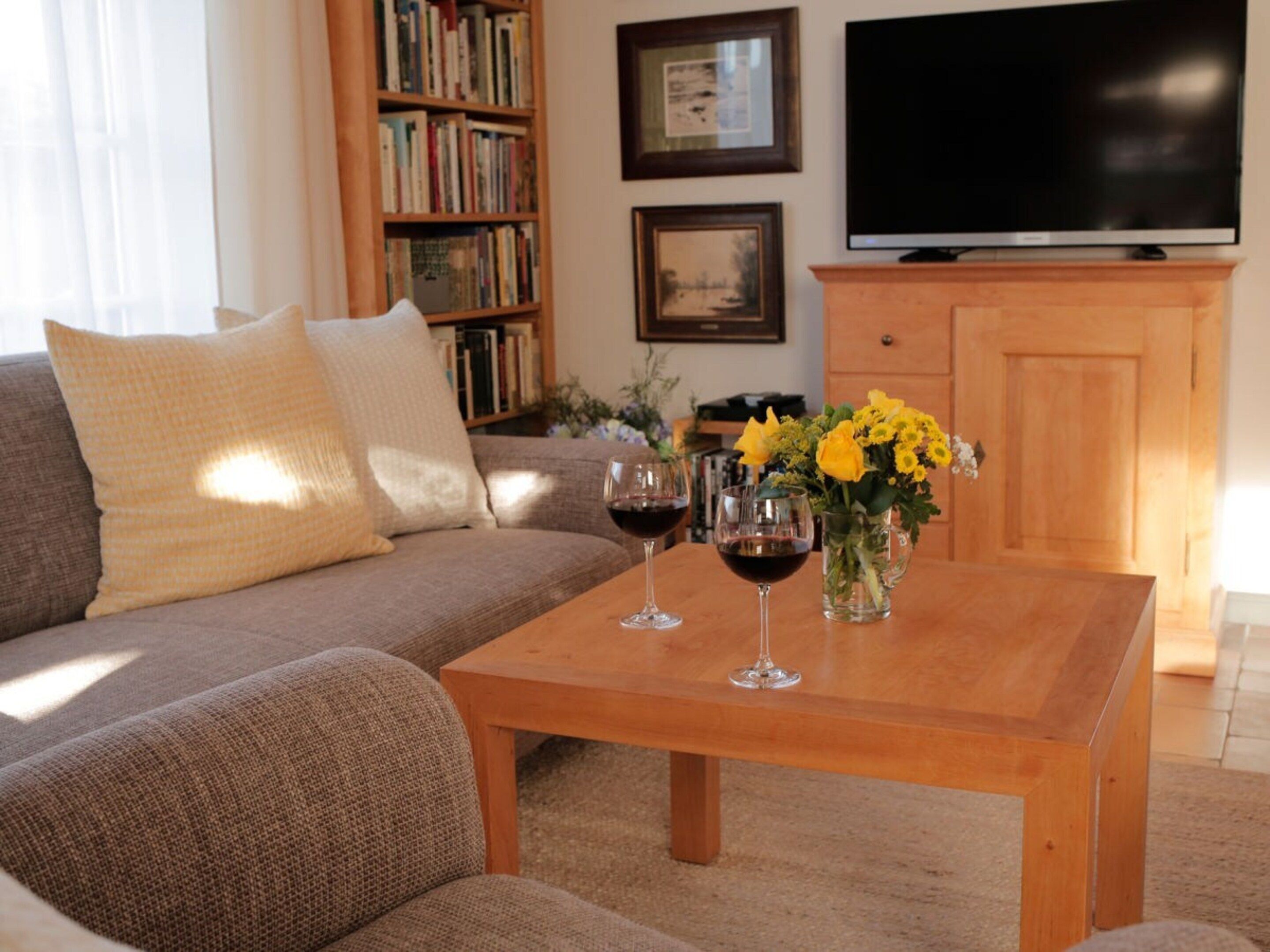 Un salon comprenant un canapé, une table basse avec des verres à vin et des fleurs, une bibliothèque, un téléviseur à écran plat et un meuble en bois.