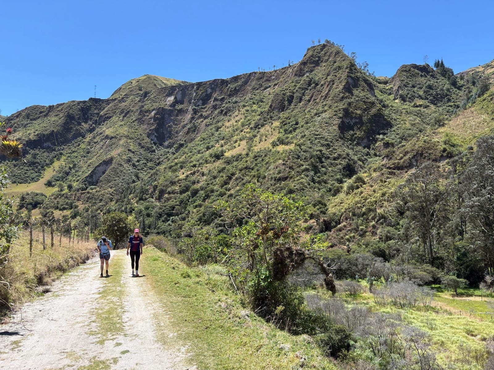 Un sendero de tierra con dos personas caminando, rodeado de colinas verdes y vegetación escasa.