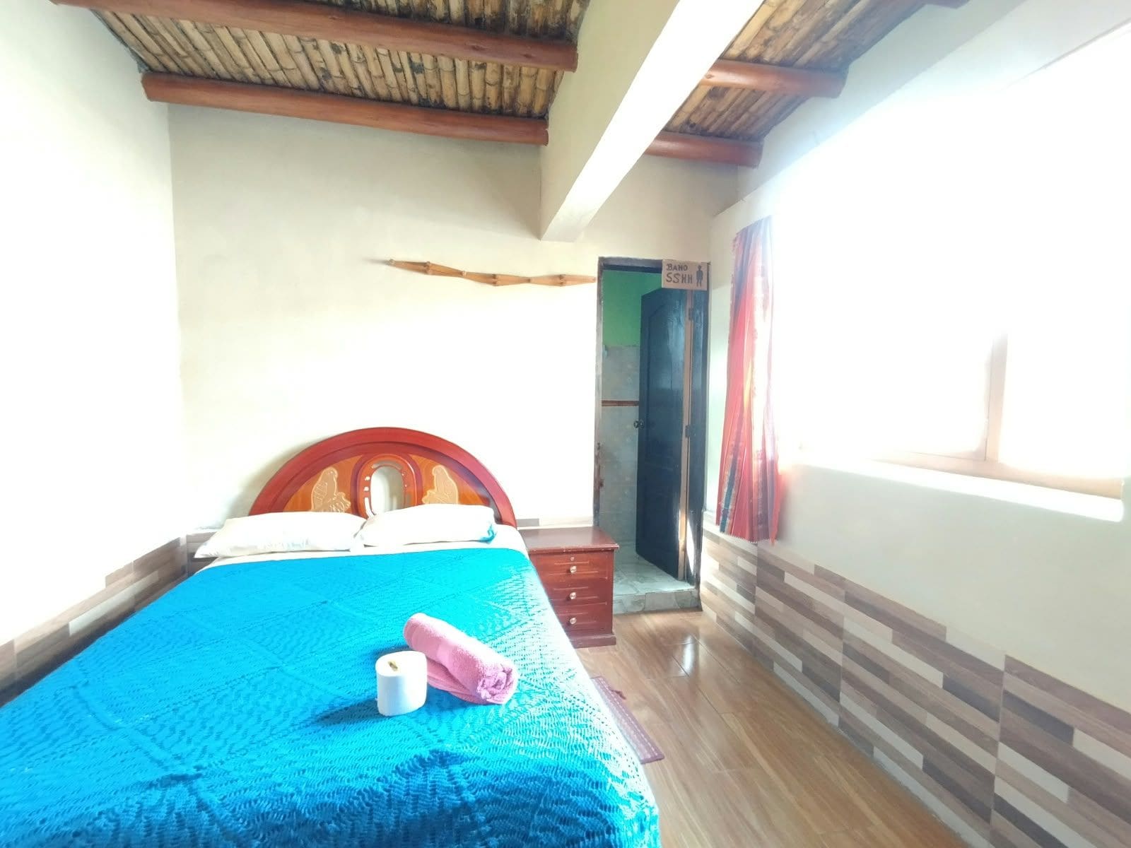 Una habitación con una cama, cabecera de madera, colcha azul, suelo de madera, una puerta y una ventana.