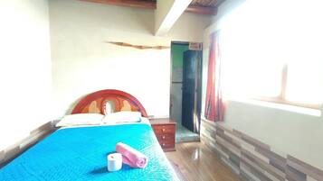 Una habitación con una cama, cabecera de madera, colcha azul, suelo de madera, una puerta y una ventana.