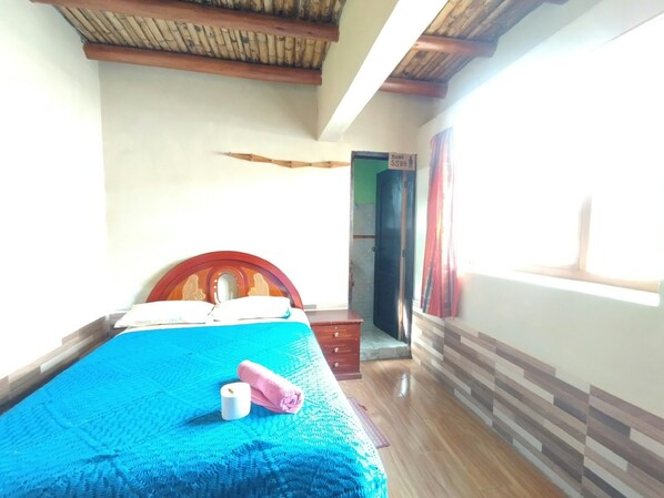 A room with a bed, a wooden headboard, a blue bedspread, a wooden floor, a door, and a window.